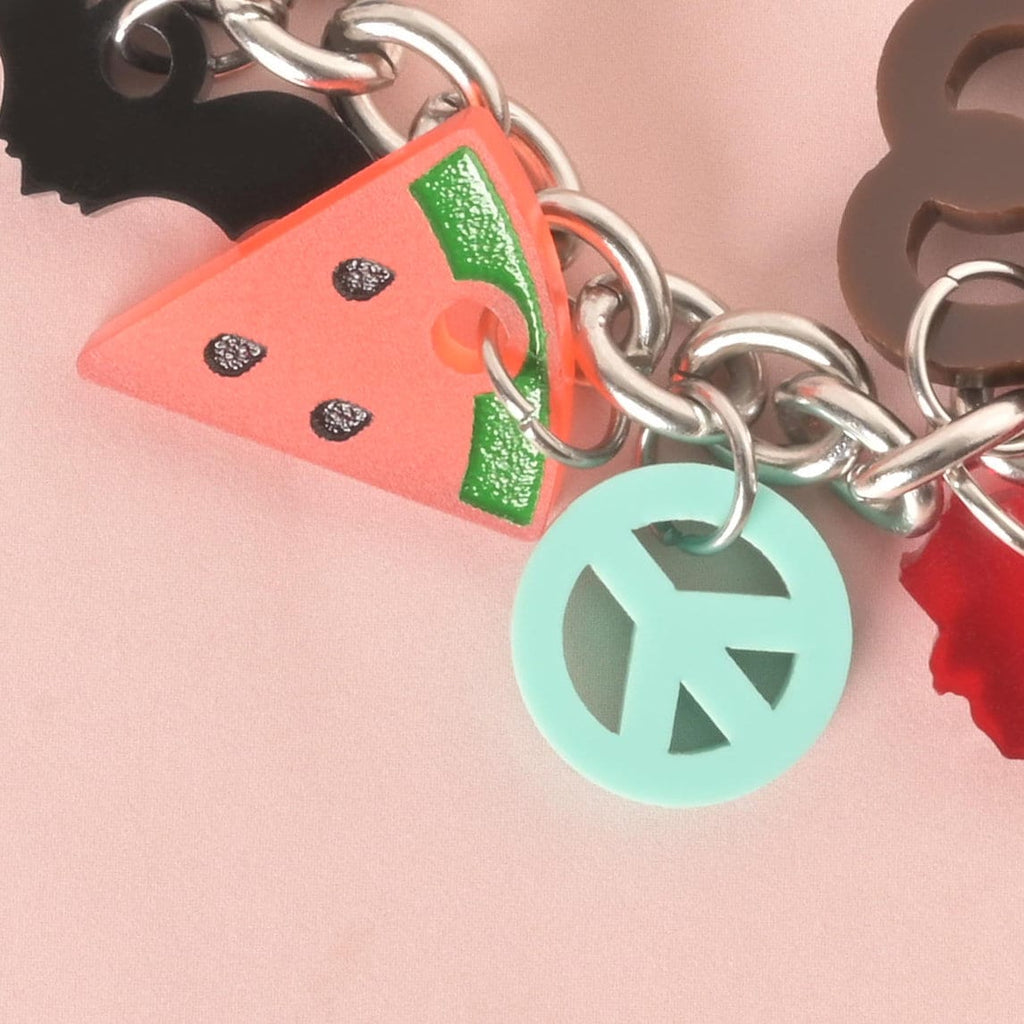 A close-up shot of a thick silver chain with several charms attached. The charms are a pink and green watermelon slice with black seeds, a translucent blue-green peace sign, and a black bat shape. A brown pretzel charm is partially visible. The charms and chain are displayed on a light pink background.