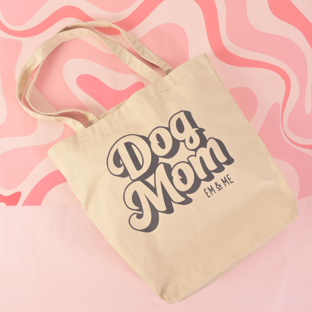A product photo showing a light-colored canvas tote bag with two handles. The front of the bag is printed with a retro-style graphic that reads "Dog Mom" and "EM & ME." The bag is positioned on a pink background with a swirling, wavy pattern.