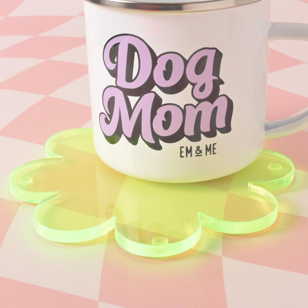 A close-up shot of a white mug that says "Dog Mom" in purple letters, sitting on a translucent fluorescent green, flower-shaped coaster. The background is a pink and white checkered pattern.