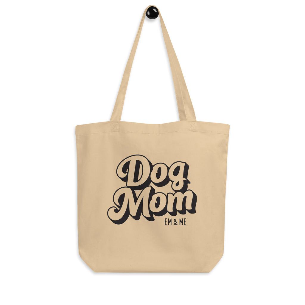 A close-up of a beige organic cotton tote bag hanging from a hook. The bag has the words "Dog Mom" printed on it in a black and white groovy font, with "EM & ME" in smaller letters below. The background is a solid light gray color.