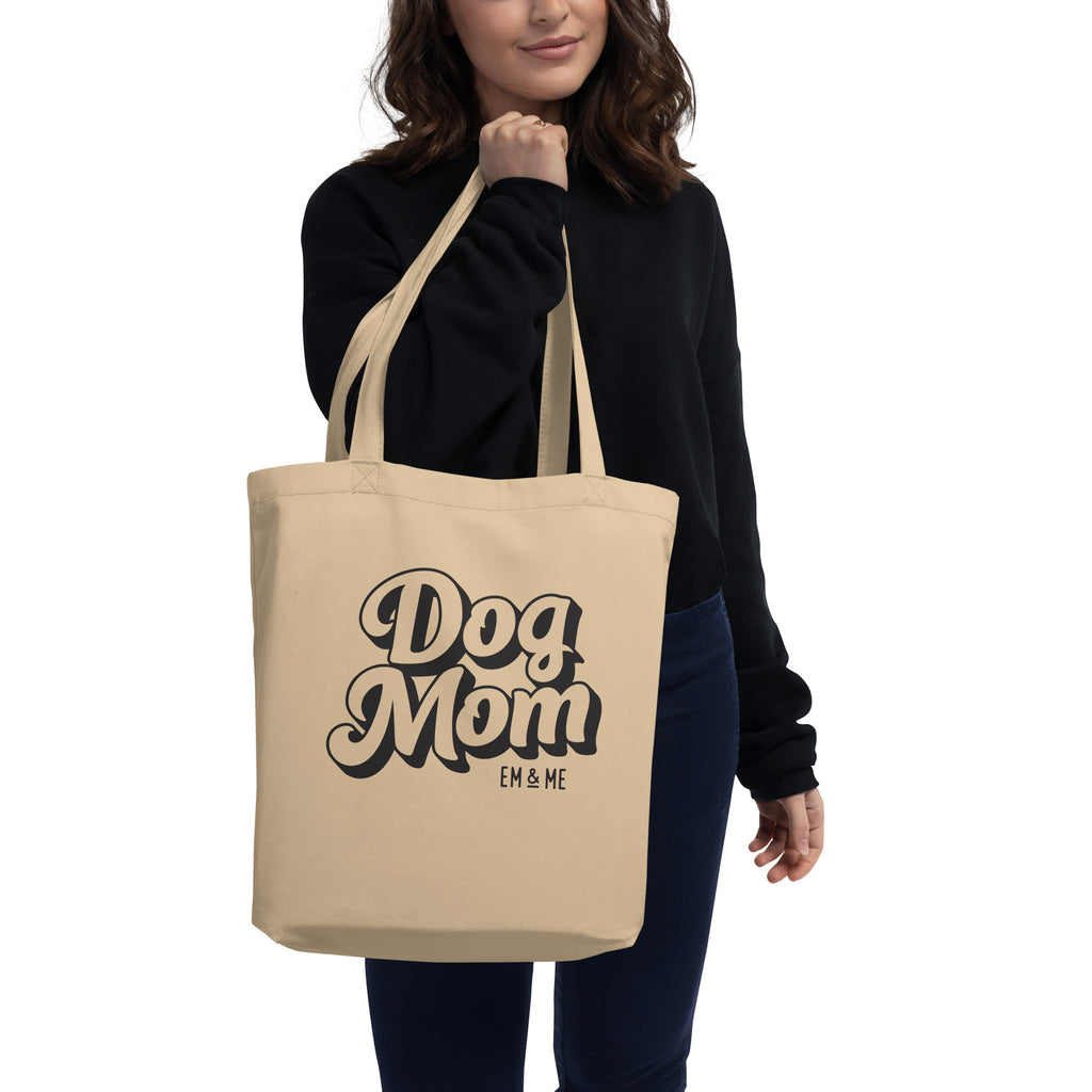 A woman wearing a black sweater holds a beige organic cotton tote bag that says "Dog Mom" in a black and white groovy font. The woman is smiling, and the bag hangs at her side. The background is a solid light gray color.
