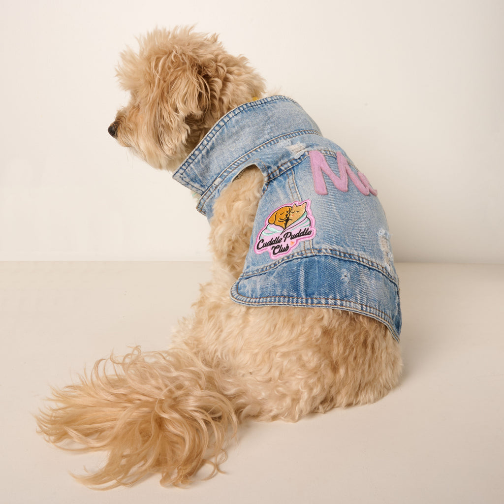 A fluffy, light-colored dog sits facing away from the camera, wearing a stylish blue denim vest. The back of the vest features a pink "ME" appliqué and a colorful patch that says "Cuddle Puddle Club." The dog is on a plain light beige background.