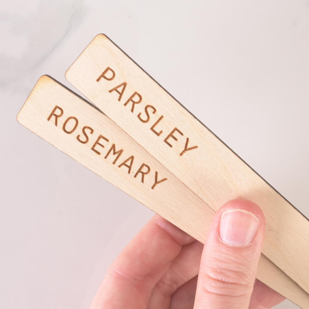 Close-up of two wooden herb markers labeled Parsley and Rosemary, engraved in uppercase letters, held in hand for gardening use.