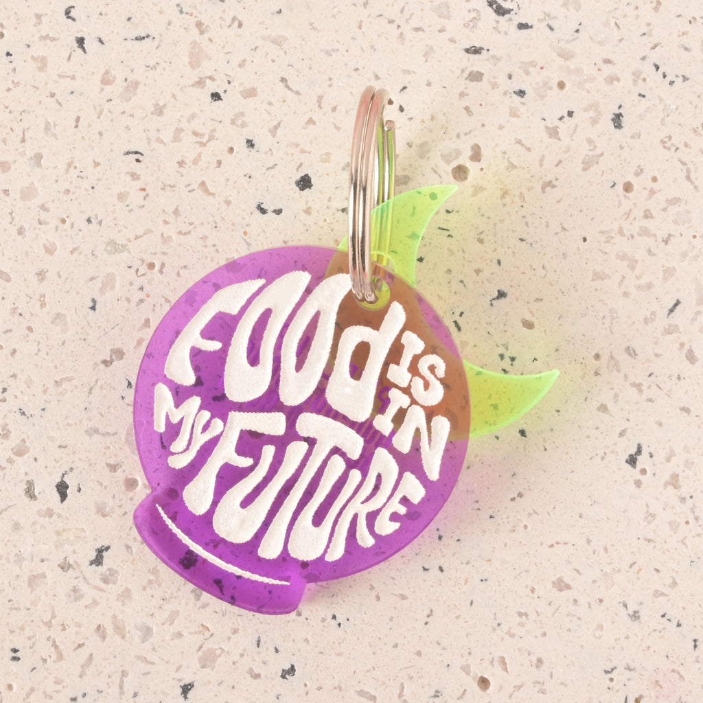 A close-up of a purple orb-shaped pet tag that says "FOOD IS IN MY FUTURE" in a white groovy font. A translucent yellow moon charm is visible behind the tag. The tag is attached to a silver ring and is resting on a light-colored, speckled surface.