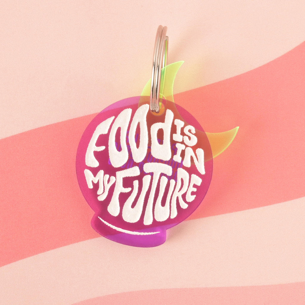 A purple orb-shaped pet tag that says "FOOD IS IN MY FUTURE" in a white groovy font. A translucent yellow moon charm is visible behind the tag. The tag is attached to a silver ring and is resting on a pink background with a wavy design.