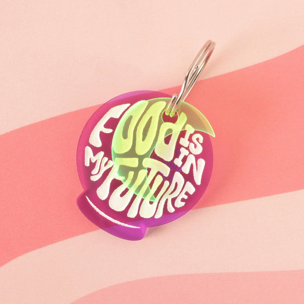 A purple orb-shaped pet tag that says "FOOD IS IN MY FUTURE" in a white groovy font. A translucent green moon charm is visible behind the tag. The tag is attached to a silver ring and is resting on a pink background with a wavy design.