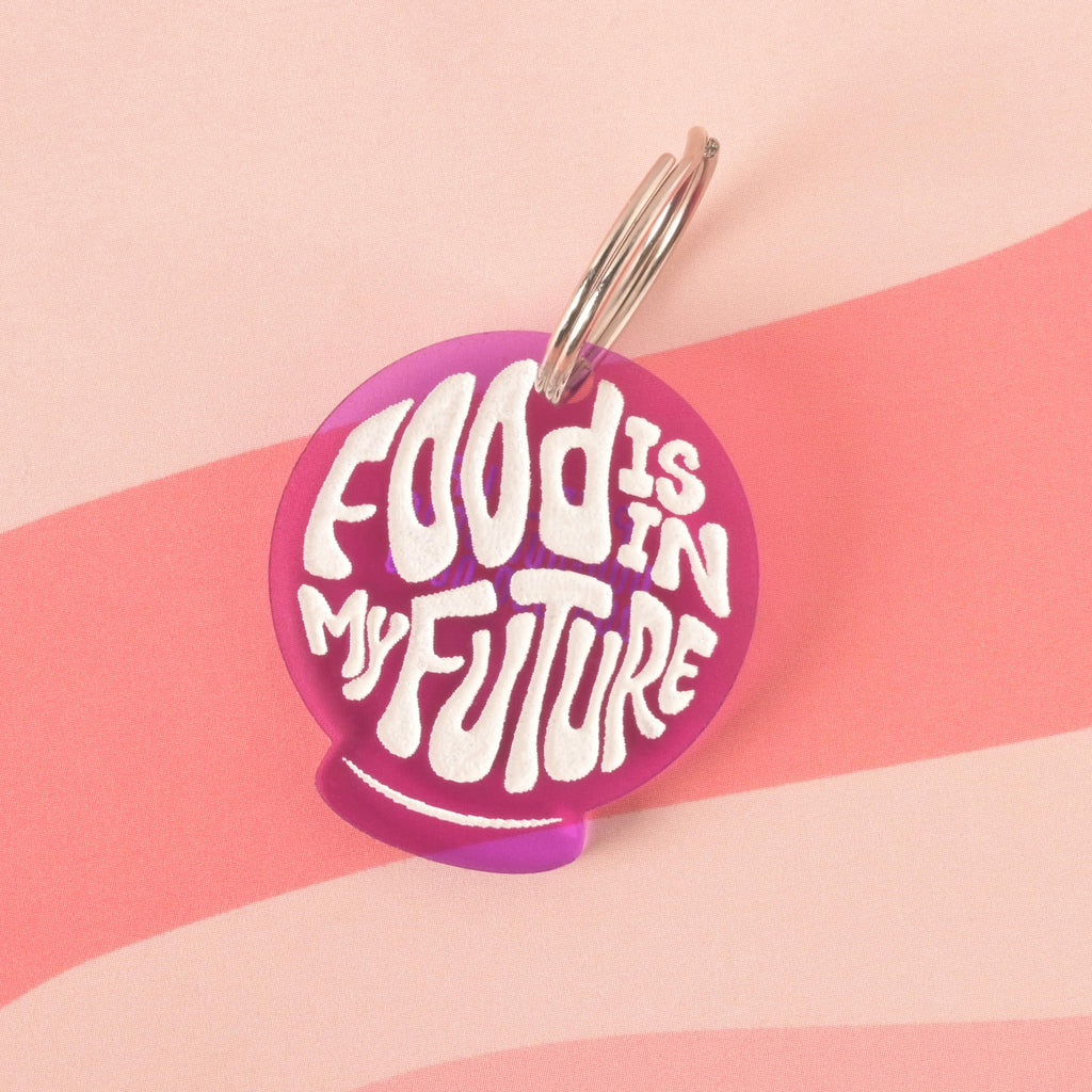 A purple orb-shaped pet tag that says "FOOD IS IN MY FUTURE" in a white groovy font. The tag is attached to a silver ring and is resting on a pink background with a wavy design.