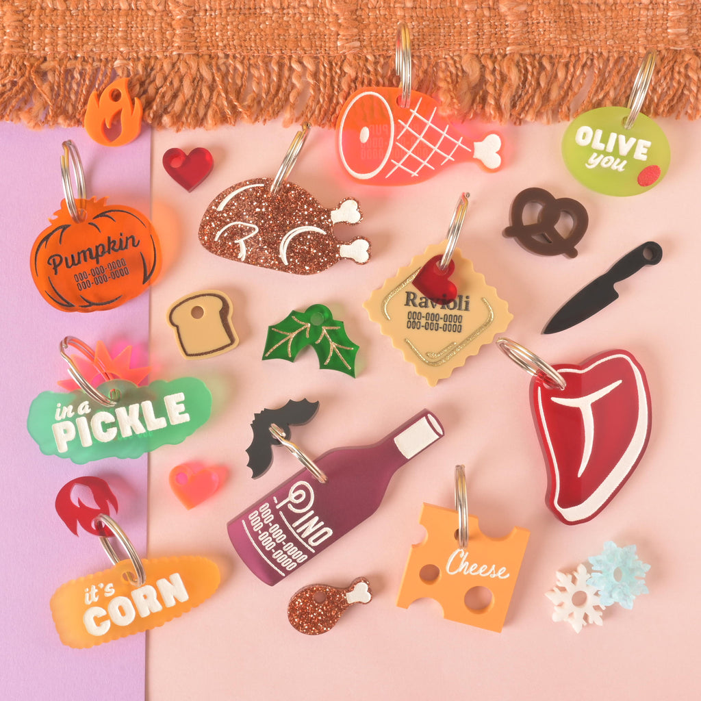 Colorful acrylic pet ID tags in playful shapes like pumpkin, steak, wine bottle, cheese, corn, ravioli, ham, pickle, pretzel, and snowflake, customized with pet names and phone numbers.