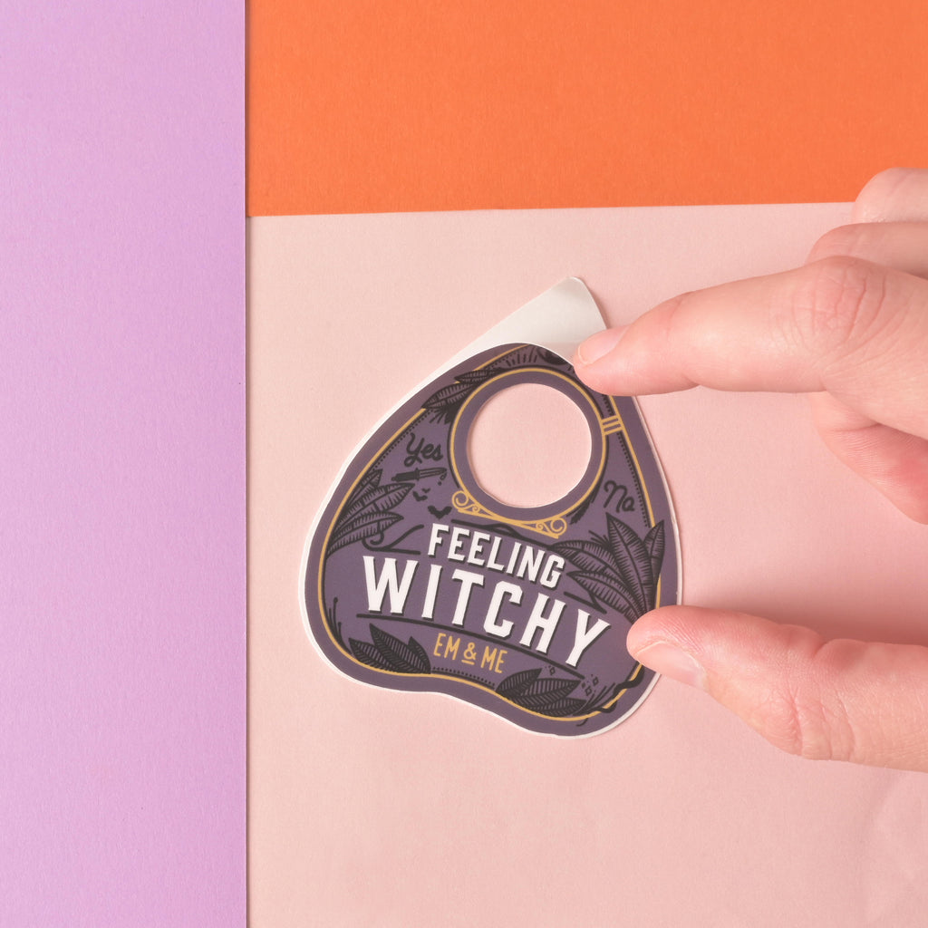 hands peeling the edge of an ouija planchet-shaped die cut sticker that reads feeling witchy" on color blocked background.