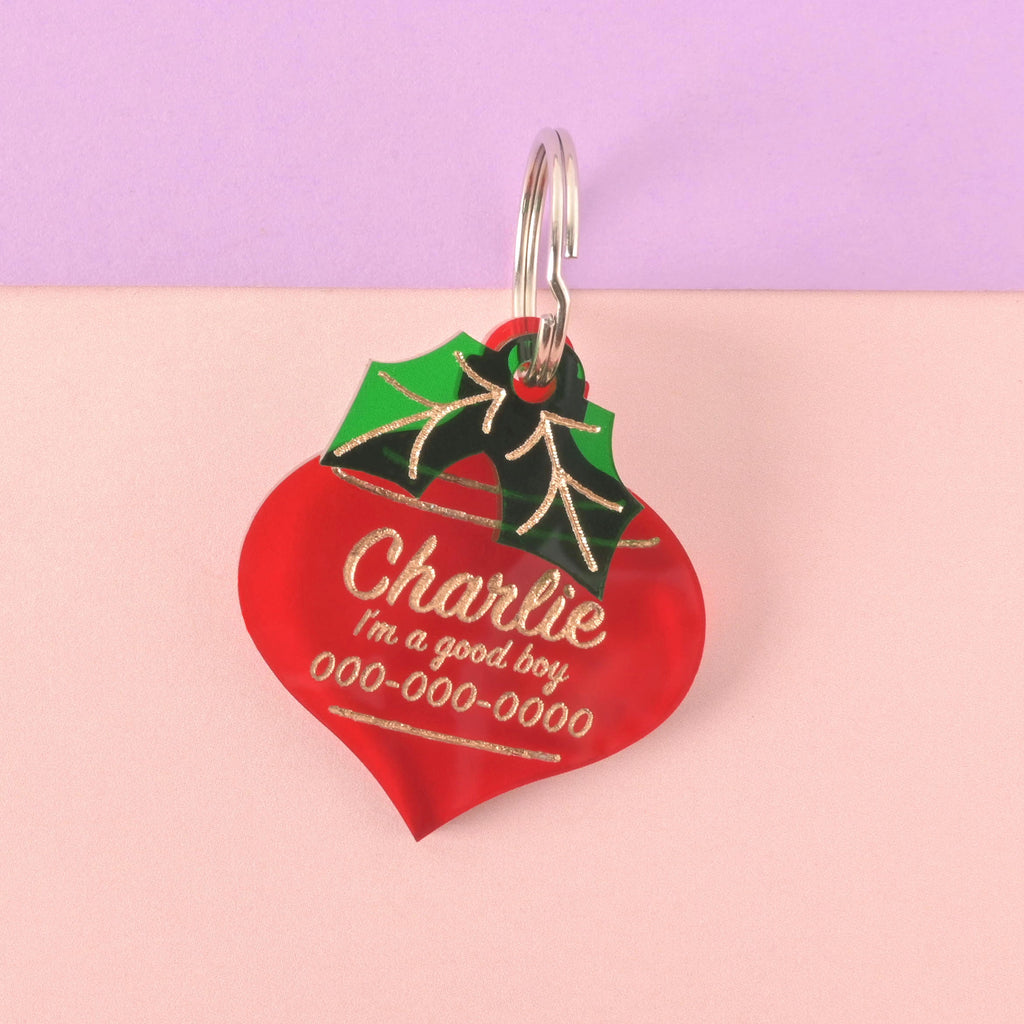 Close-up of a large translucent red acrylic pet ID tag shaped like a Christmas ornament, featuring gold engraved personalization for 'Charlie' and safety information. A green acrylic holly leaf charm with gold details is layered on top. The tag is displayed on a color-blocked background of lavender and light pink.