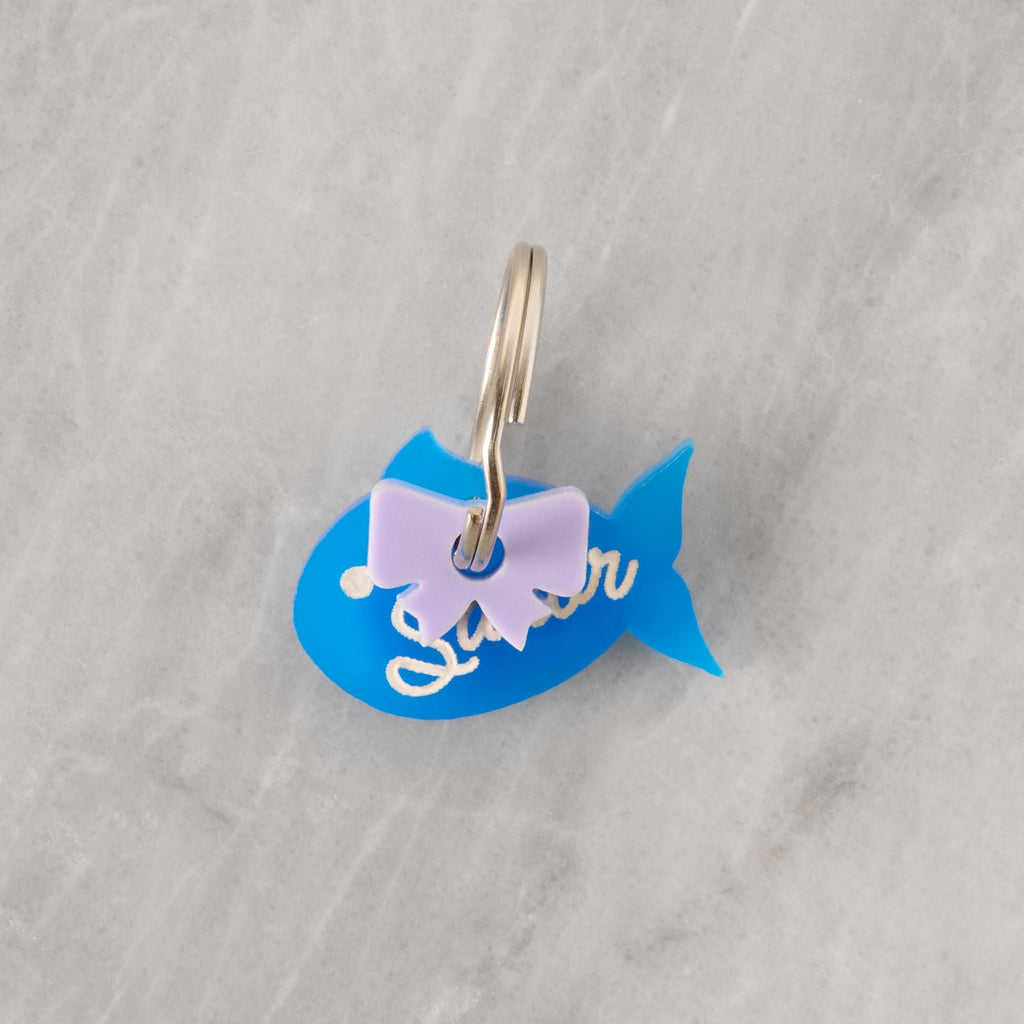 This is a photo of a keychain shaped like a blue fish. It has a small lavender bow attached near the top, and the word "Sailor" is written on the fish in white cursive lettering. The keychain is attached to a metal ring and placed on a gray marble-like surface.