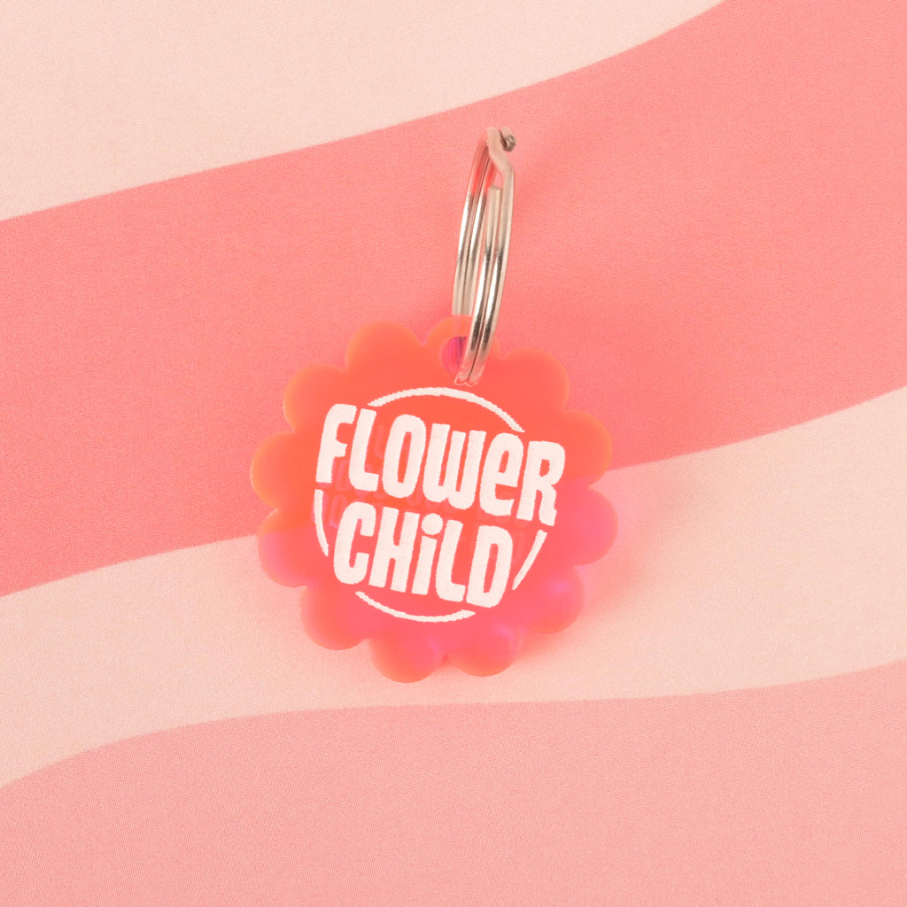  A Fluorescent pink flower-shaped pet tag that says "FLOWER CHILD" in white letters. The tag is attached to a silver ring and is resting on a pink background with a wavy design.