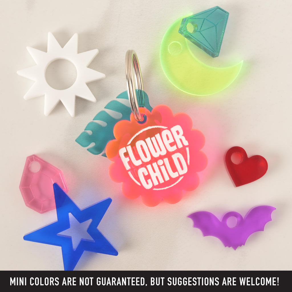 A collection of pet tags and charms scattered on a white surface. The central piece is a pink and orange "FLOWER CHILD" tag with a light blue leaf charm. Surrounding it are various charms, including a white sunburst, a pink gemstone, a blue star, a red heart, a purple bat, and a translucent yellow moon with a green and teal diamond. The image includes the text "MINI COLORS ARE NOT GUARANTEED, BUT SUGGESTIONS ARE WELCOME!"