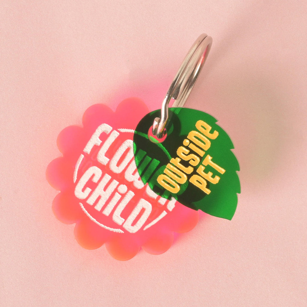 A custom, pink, flower-shaped pet ID tag with "FLOWER CHILD" engraved in white font. A small, dark green leaf-shaped charm with the text "OUTSIDE PET" in yellow is attached to the silver keyring. The tags are displayed on a solid pink background.