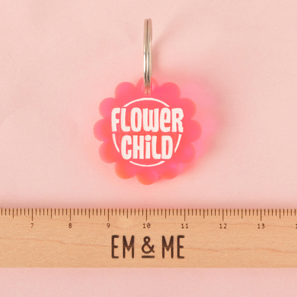 A pink flower-shaped pet tag that says "FLOWER CHILD" in white letters. A ruler with the "EM & ME" logo is placed below the tag to show its size. The tag is attached to a silver ring and is resting on a solid pink background.
