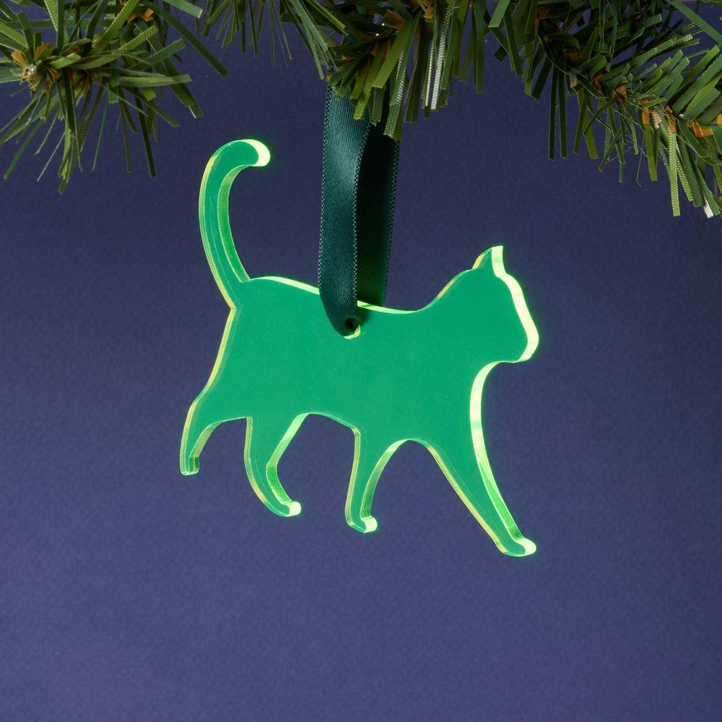 A laser-cut fluorescent neon green acrylic cat silhouette ornament, hanging by a dark green ribbon from a pine branch. This vibrant pet décor is set against a dark blue background.