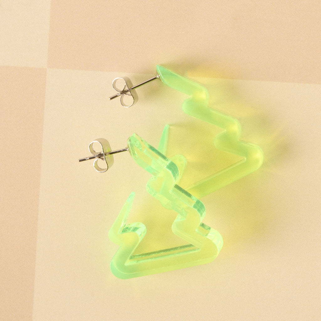 A pair of unique holiday earrings shaped like Christmas trees or zig-zag hoops in fluorescent neon yellow-green acrylic. The earrings are lightweight and displayed against a beige and pink background.