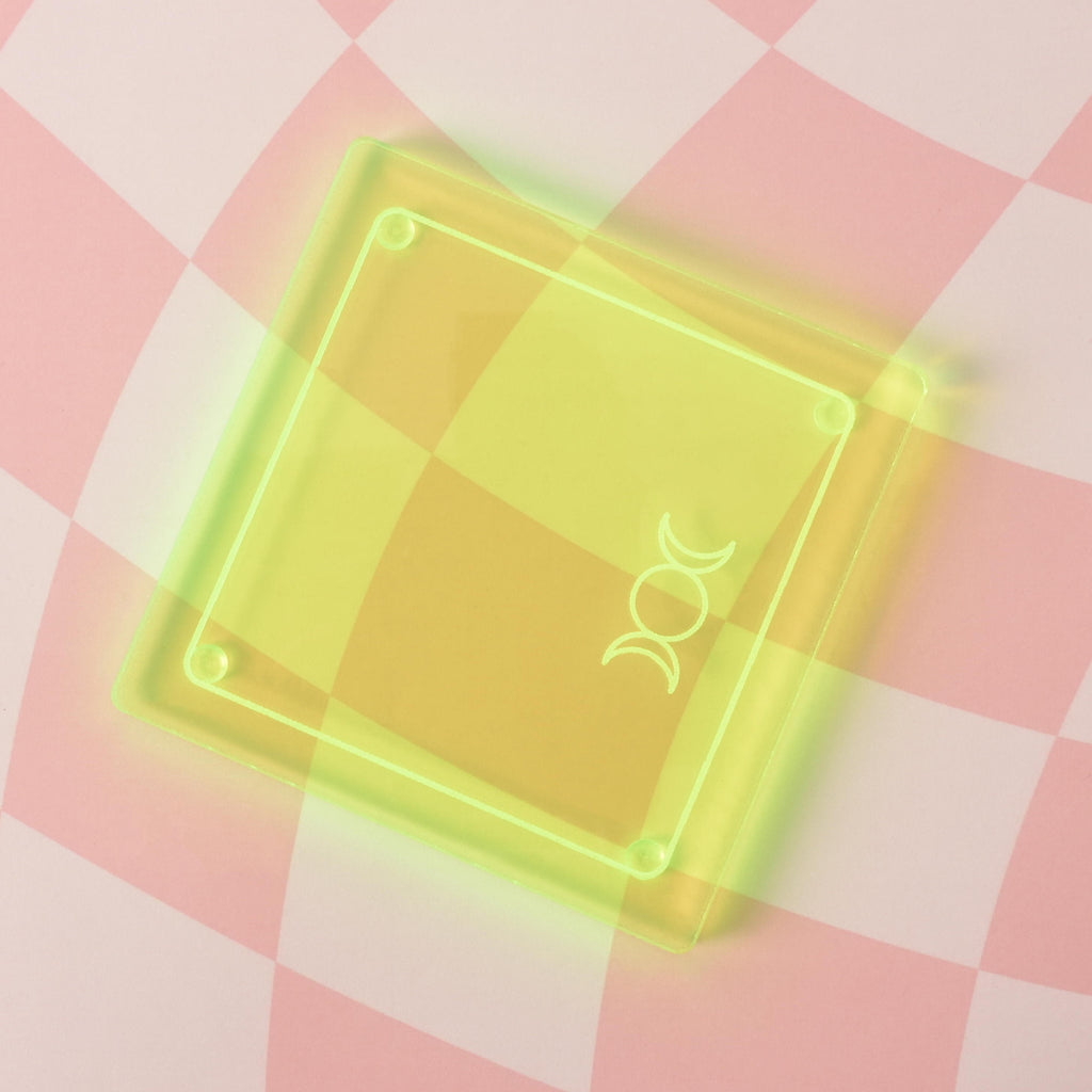 A single square-shaped, translucent yellow coaster is shown on a pink and white checkered background. The coaster has a gold-etched triple moon symbol.
