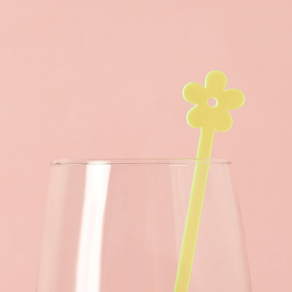 A single Fluorescent Green flower-shaped cocktail stirrer is placed inside a clear glass. The background is a solid pink color.