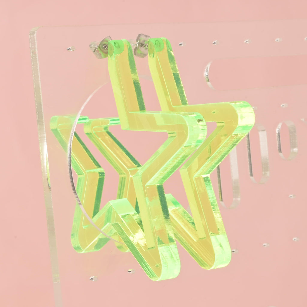  pair of large, star-shaped hoop earrings made from neon green iridescent acrylic. The earrings have silver posts and are displayed hanging from a clear earring holder against a solid pink background. The iridescent material reflects a yellowish-green glow.