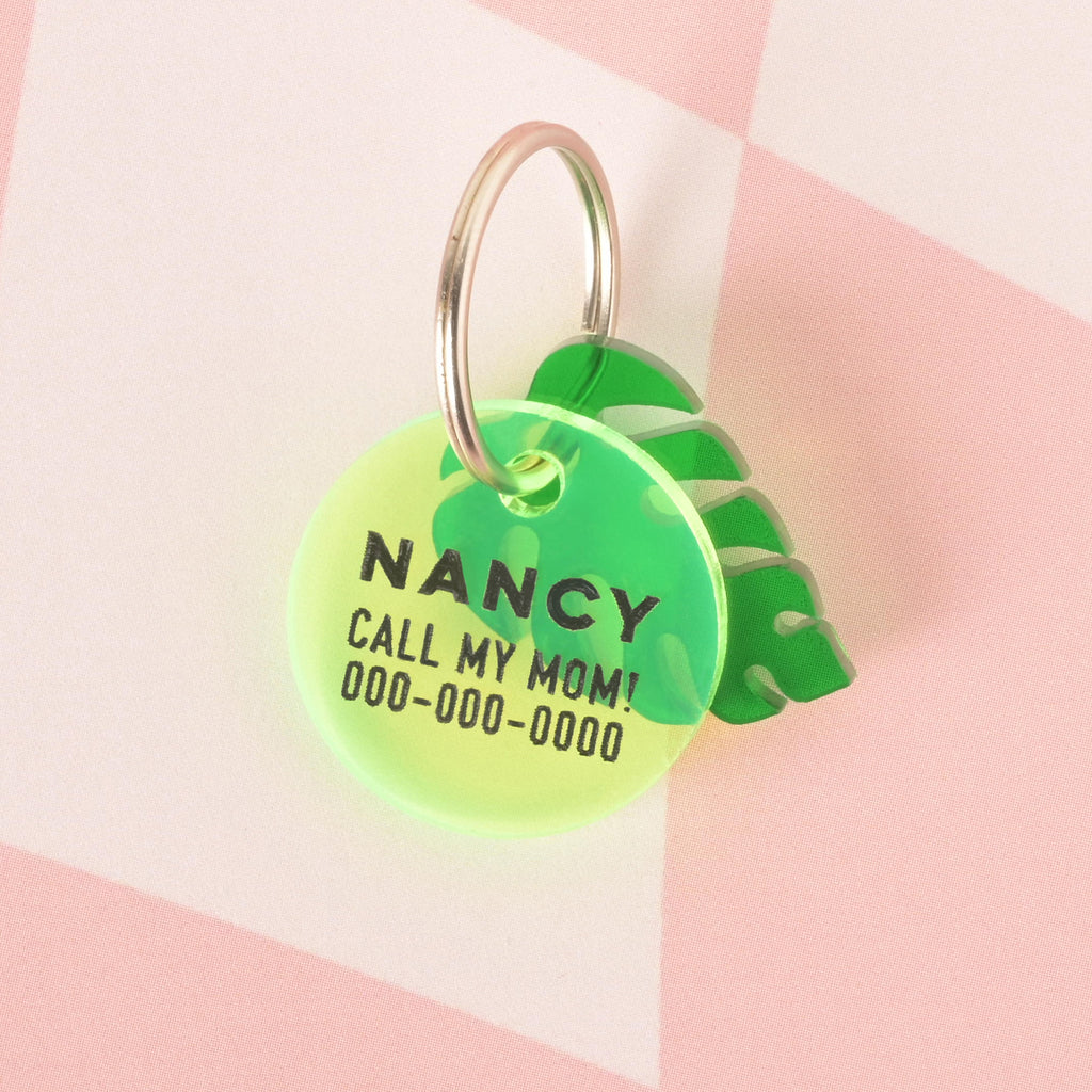 A close-up product photo of a round, neon yellow pet tag with a green gradient. A green monstera leaf charm is attached to the tag, which is engraved with the name "NANCY" and the phrase "CALL MY MOM!" in black text. A silver split key ring is also shown.