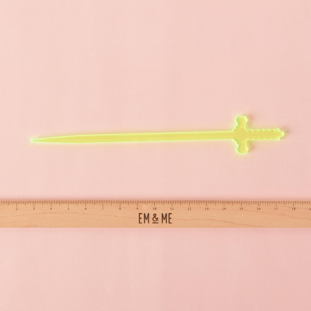 A single translucent Fluorescent Green, sword-shaped cocktail stirrer is shown on a solid pink background. A ruler with the "EM & ME" logo is placed below the stirrer to show its size.