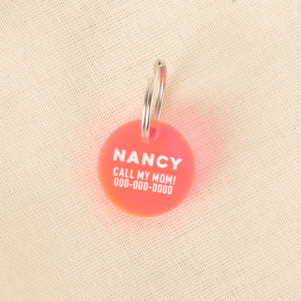 A close-up product photo of a round, red and orange gradient pet tag with the name "NANCY" and the funny phrase "CALL MY MOM!" engraved on it. The tag is shown with a silver split key ring on a light-colored, woven fabric background.
