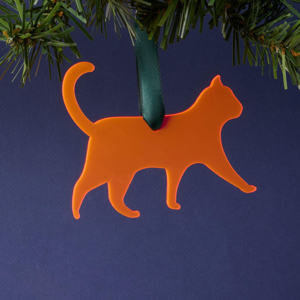 A laser-cut fluorescent pink acrylic cat silhouette ornament, hanging by a dark green ribbon from a pine branch. This vibrant pet décor is set against a dark blue background.