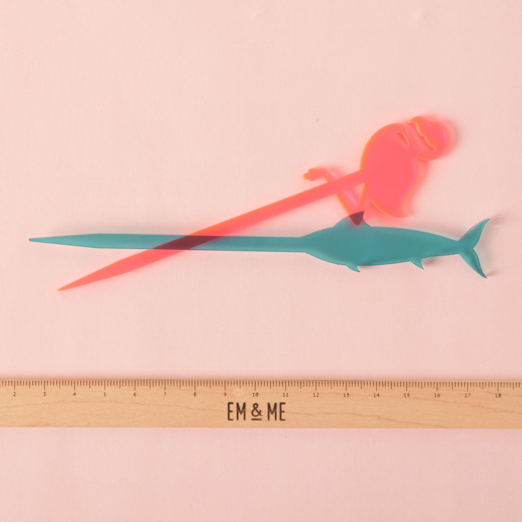 A Fluorescent Pink-shaped cocktail stirrer crosses over a teal swordfish-shaped stirrer. A ruler with the "EM & ME" logo is placed below the stirrers to show their size. The background is a solid pink color.