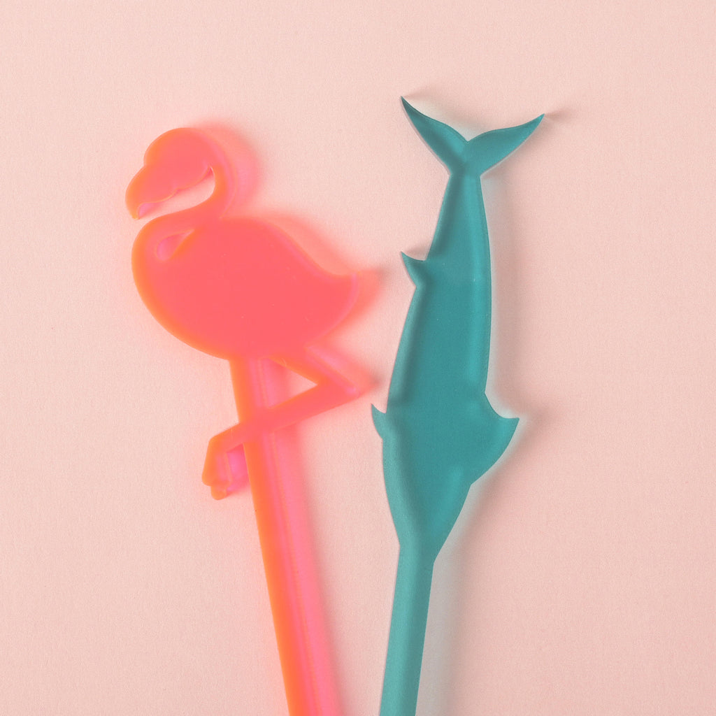A neon pink flamingo-shaped cocktail stirrer and a teal dolphin-shaped cocktail stirrer are lying side-by-side on a solid pink background.