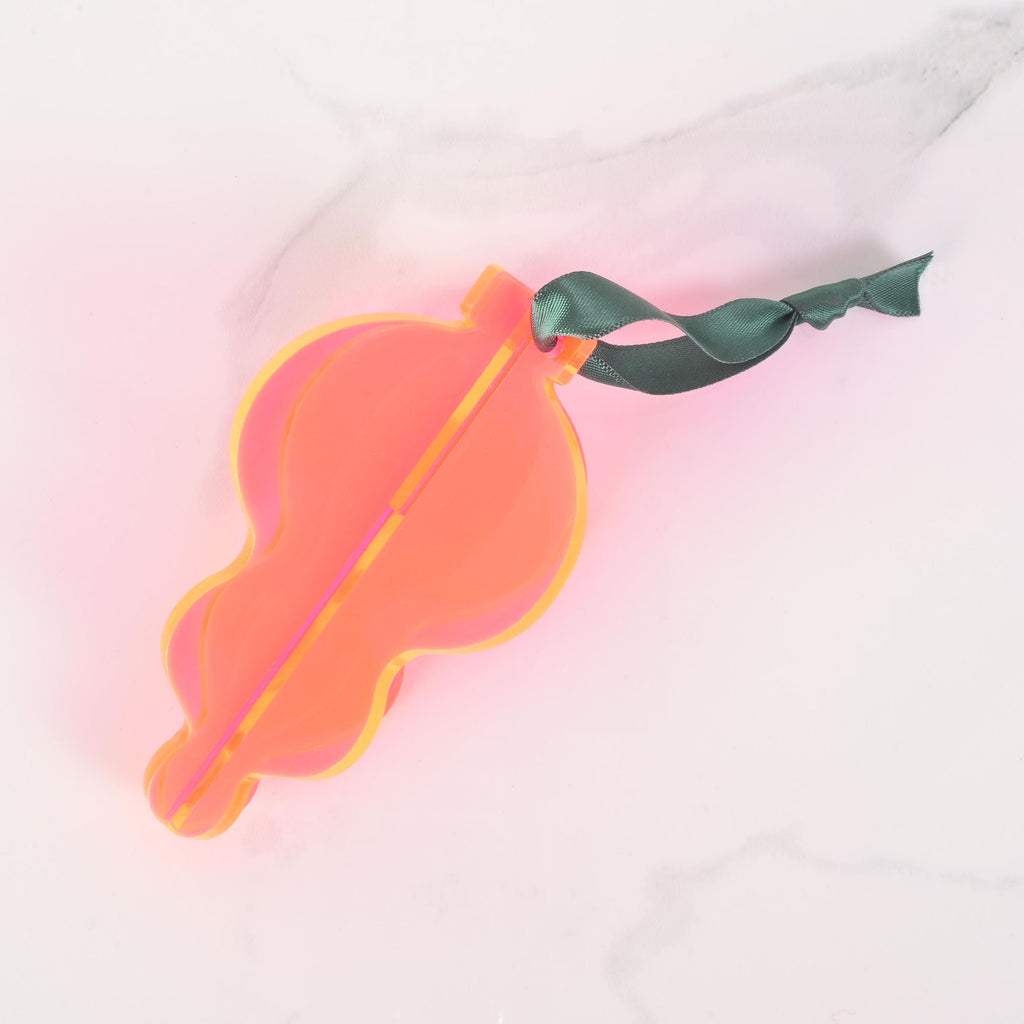 A neon pink and orange flat-pack Christmas tree ornament shaped like a classic glass ornament. The ornament is assembled and has a dark green ribbon tied to the top for hanging. The item is displayed on a white marble surface.