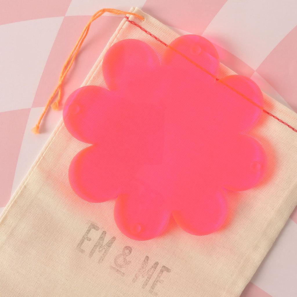 A vibrant neon pink, flower-shaped coaster is placed on a beige pouch with the "EM & ME" logo. The background is a pink and white checkered pattern.