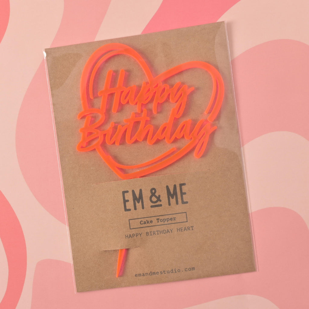 A Fluorescent Pink cake topper that says "Happy Birthday" inside a heart outline, sealed in its retail packaging. The brown cardstock packaging has "EM & ME" printed on it. The item is shown on a pink background with a wavy design.