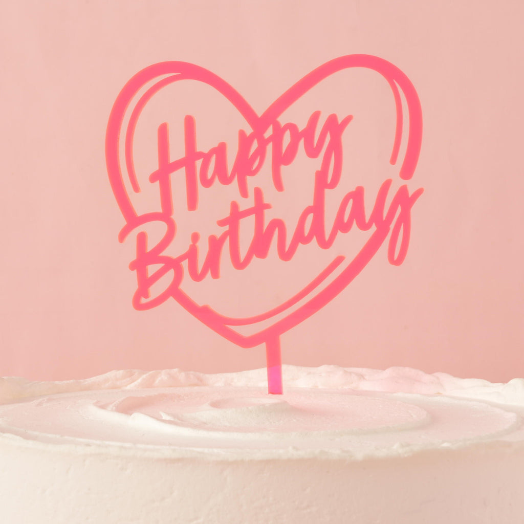 A neon pink cake topper that says "Happy Birthday" inside a heart outline, placed on top of a frosted white cake. The background is a solid pink color.