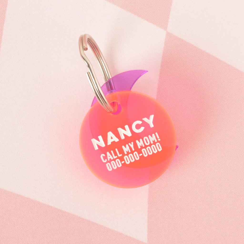 A close-up product photo of a round, red and orange gradient pet tag, also known as a Minimal Circle tag, with a purple crescent moon charm attached to the top. The tag is engraved with the name "NANCY" and the funny phrase "CALL MY MOM!" It is shown with a silver split key ring.