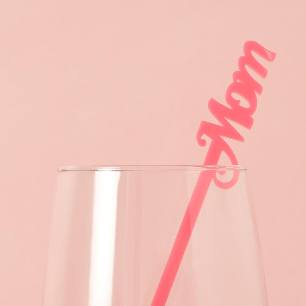 A single pink cocktail stirrer with the word "Mom" is placed inside a clear glass. The background is a solid pink color.