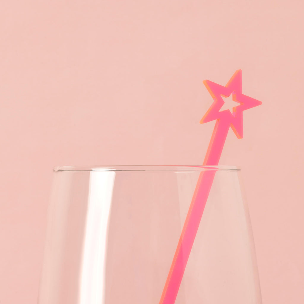 A single neon pink star-shaped cocktail stirrer is placed inside a clear glass. The background is a solid pink color.
