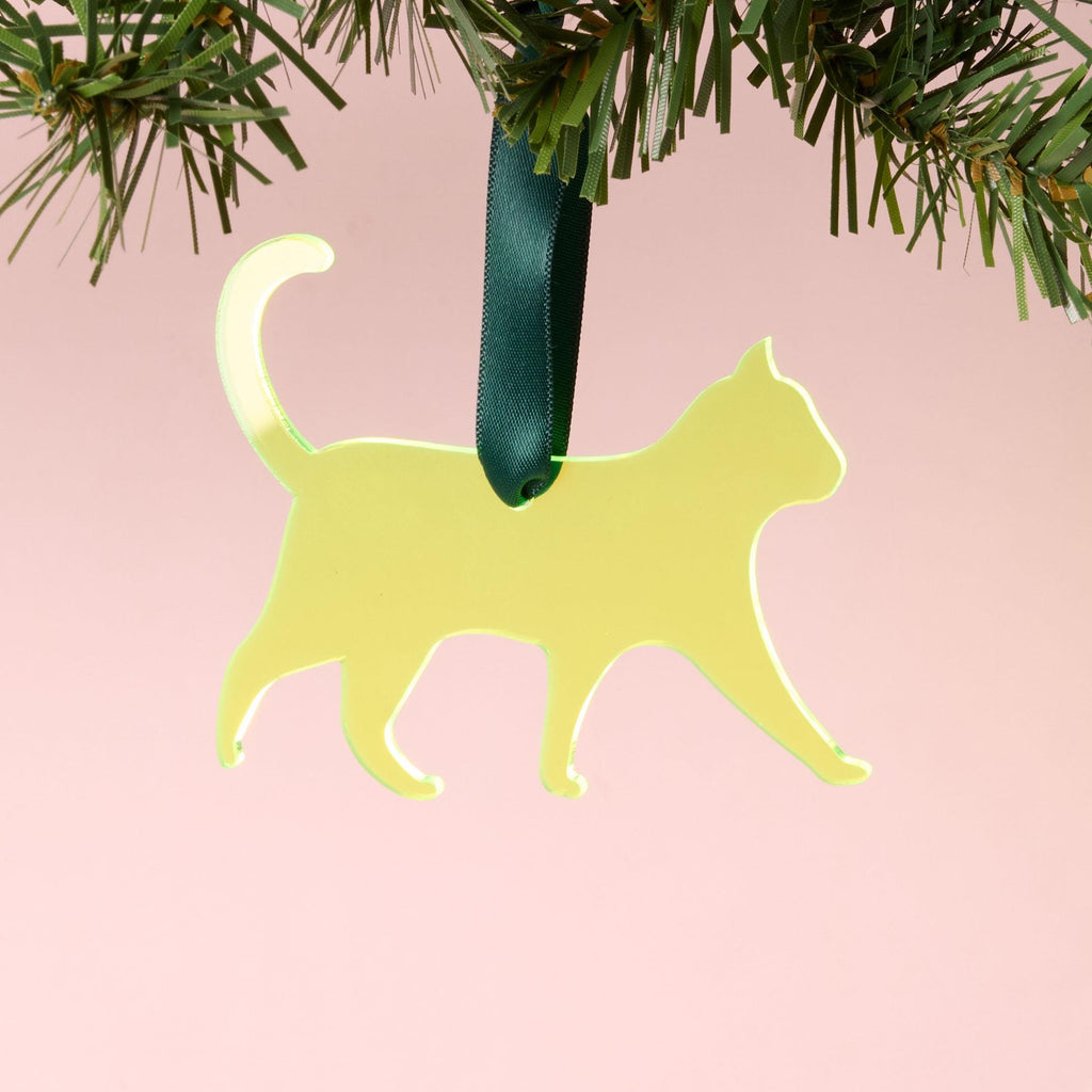 A laser-cut fluorescent neon yellow-green acrylic cat silhouette ornament, hanging by a dark green ribbon from a pine branch, set against a pink background.