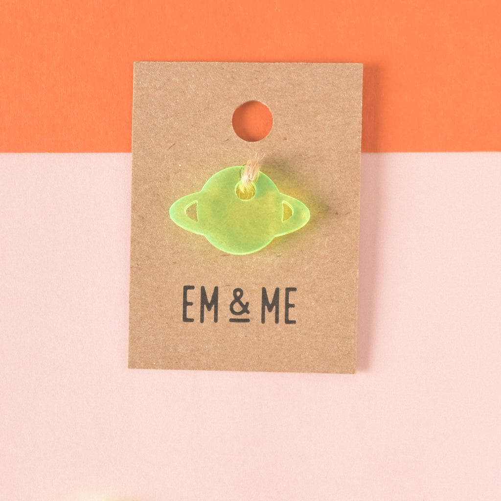 Fluorescent green Saturn-shaped mini charm packaged on brown kraft paper on color blocked background