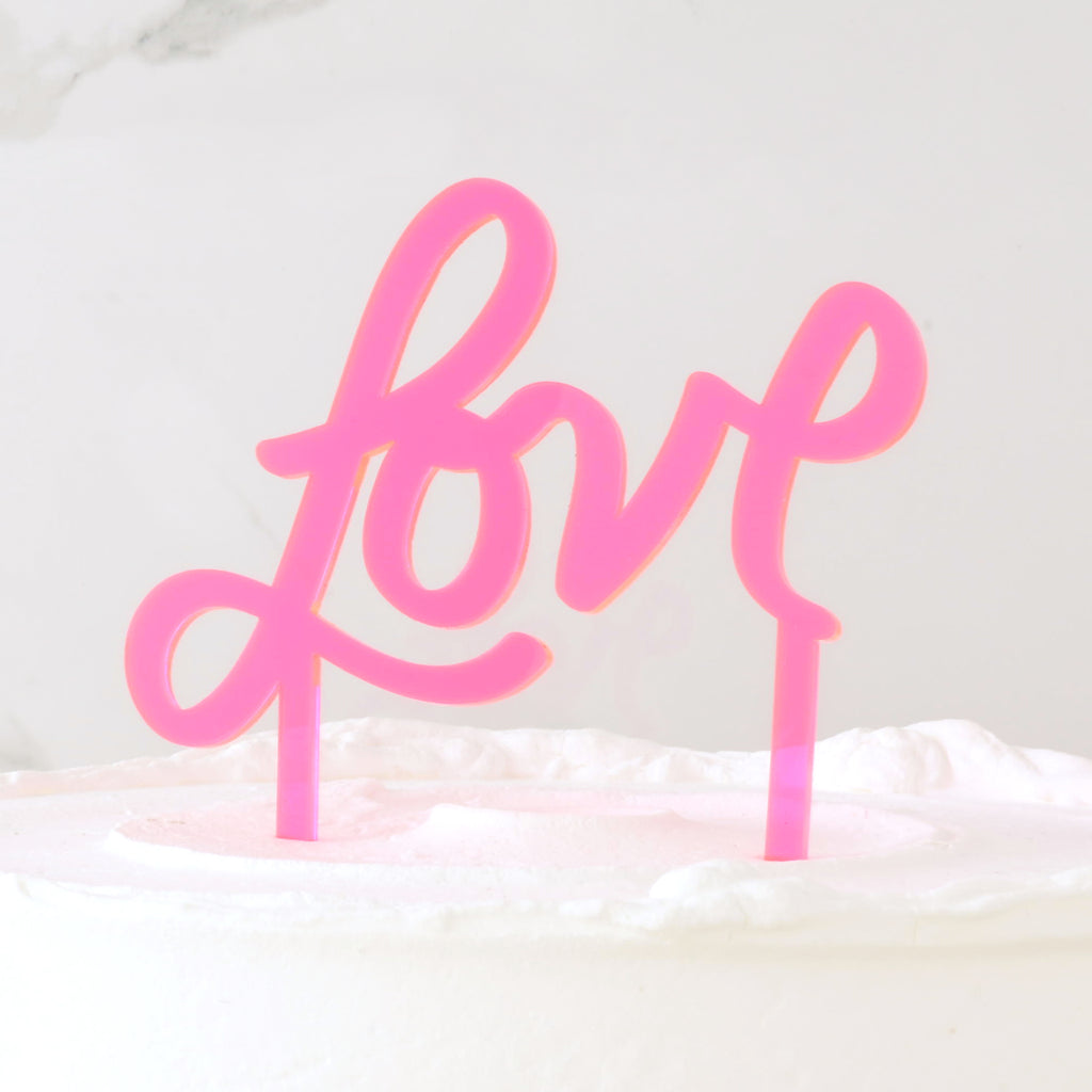 Fluorescent pink acrylic cake topper that reads "Love: in a hand drawn script font inserted on white frosted cake.
