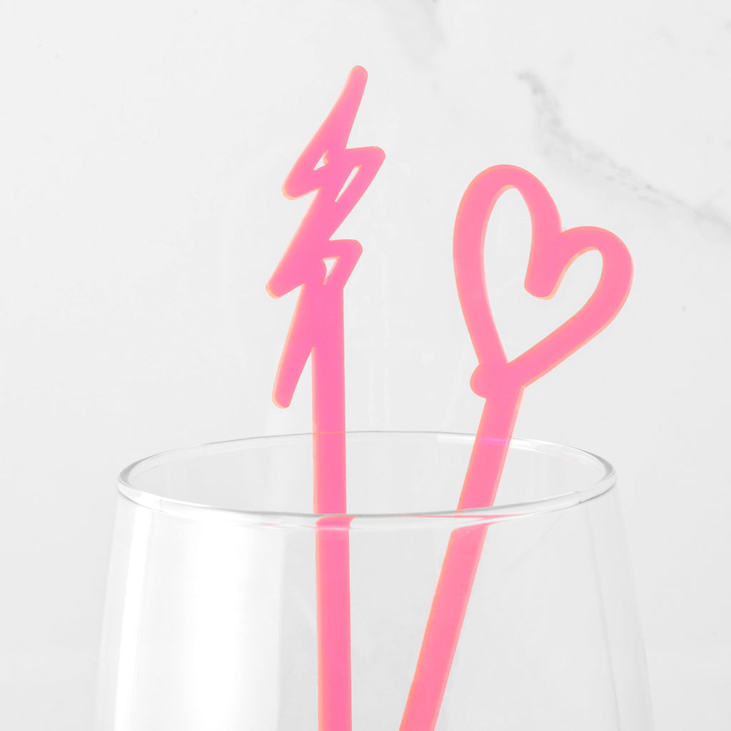 Close up of two fluorescent pink cocktail stirrers shaped like a heart and lightning bolt in a clear class on marble background
