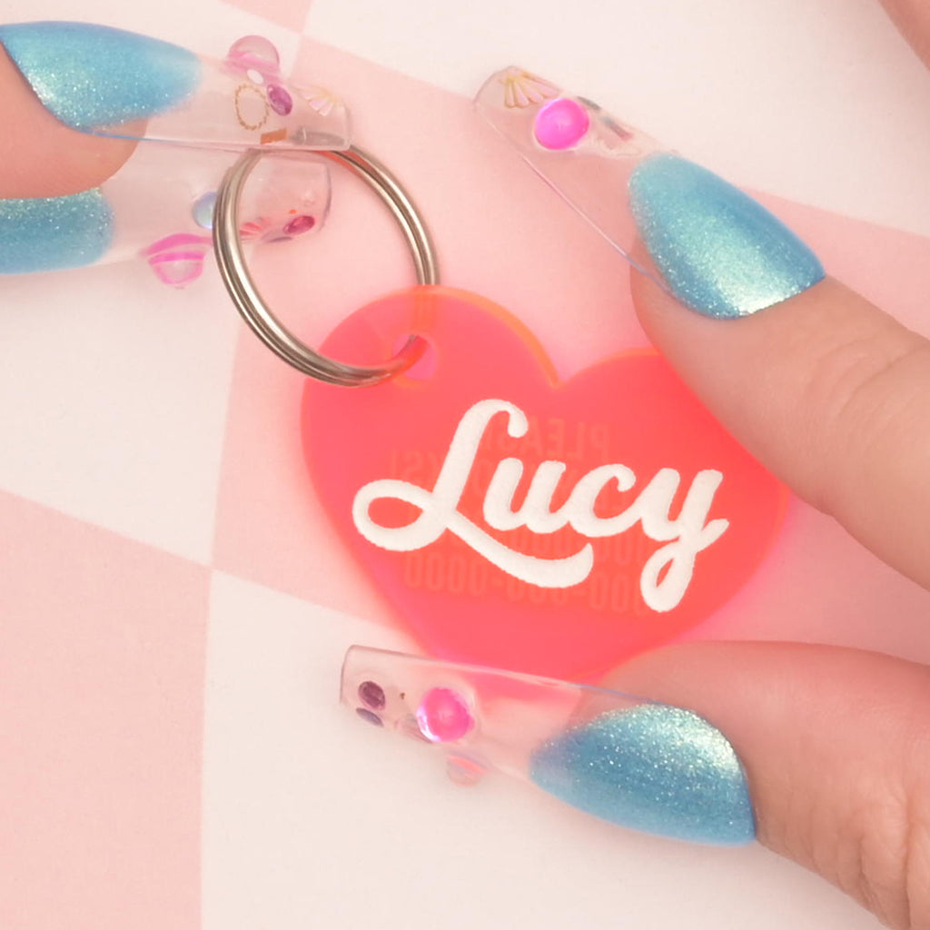 close up of hands with blue girly nails holding a fluorescent pink heart-shaped personalized pet tag on solid pink background