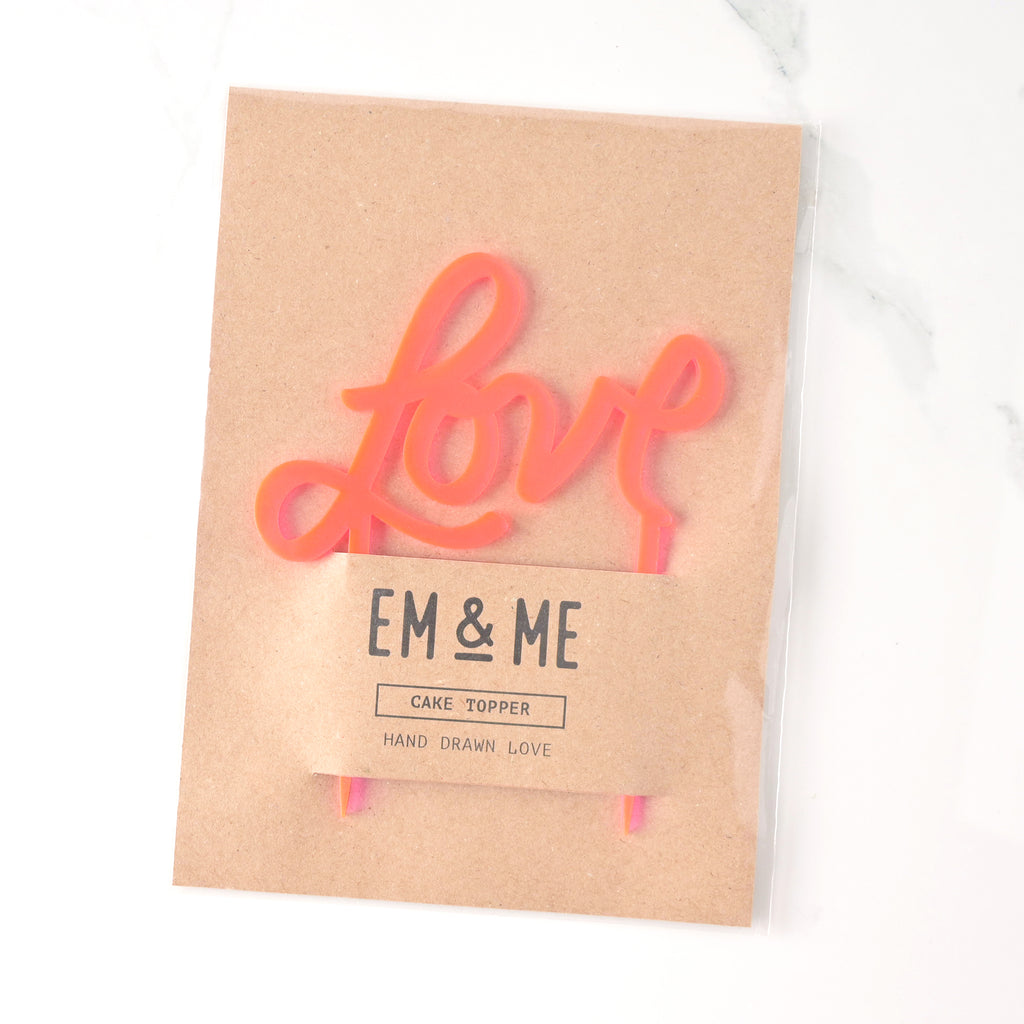 Fluorescent pink acrylic cake topper that reads "love" in a hand drawn script font packaged on brown kraft paper on marble background.
