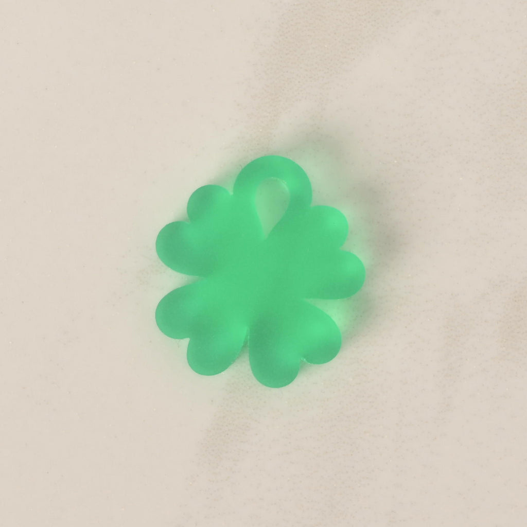 A close-up of a small, translucent green, four-leaf clover-shaped charm. The charm is displayed on a light-colored, speckled countertop or stone surface.