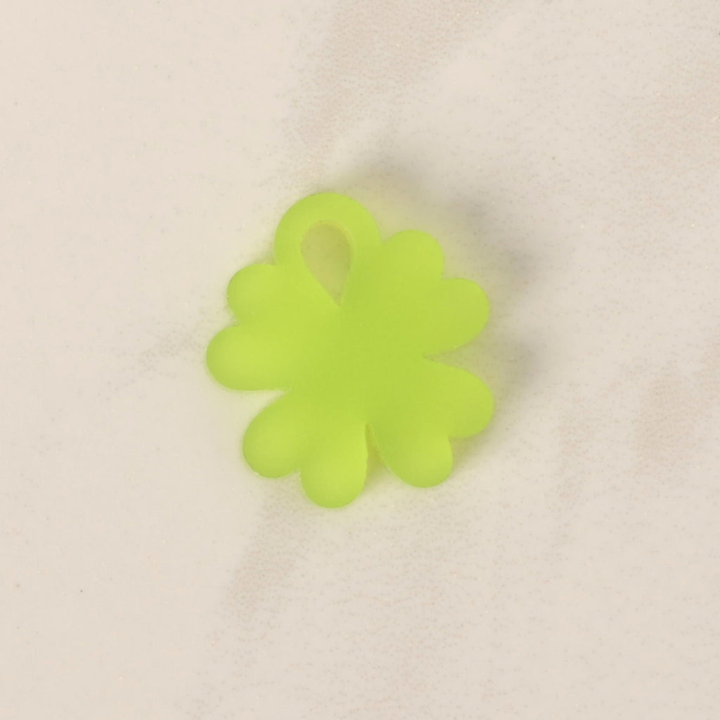 A close-up of a small, translucent neon green, four-leaf clover-shaped charm. The charm is displayed on a light-colored, speckled countertop or stone surface.