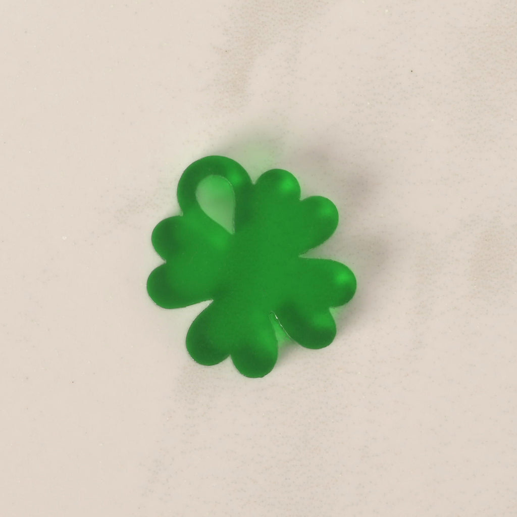 A close-up of a small, translucent green, four-leaf clover-shaped charm. The charm is displayed on a light-colored, speckled countertop or stone surface.
