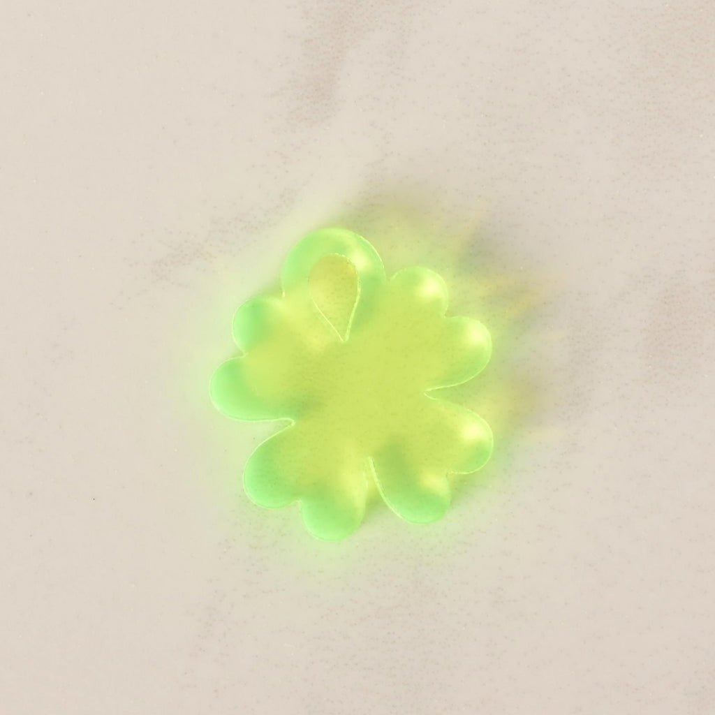 A close-up of a small, translucent neon green, four-leaf clover-shaped charm. The charm is displayed on a light-colored, speckled countertop or stone surface.