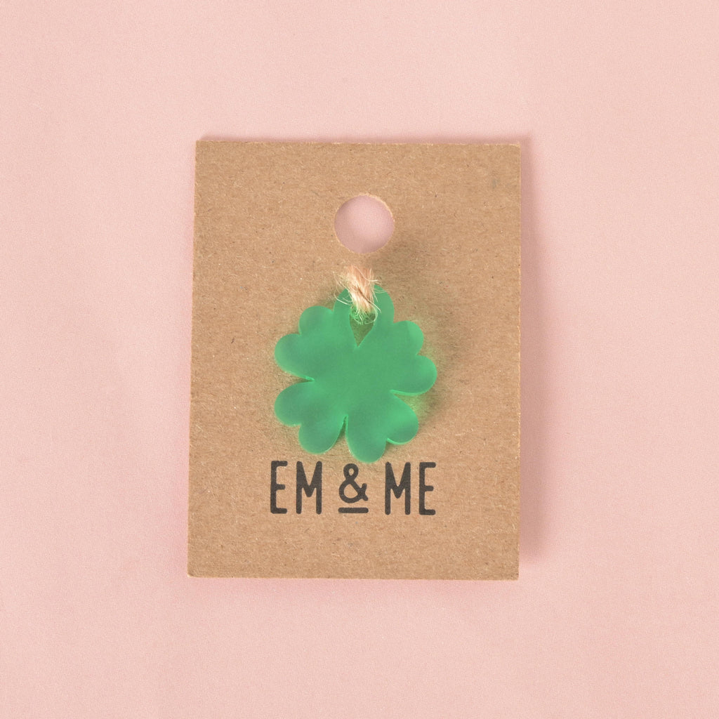 A small, translucent green, four-leaf clover-shaped charm. The charm is attached with a piece of string to a brown cardboard tag with the logo "EM & ME." The item is displayed on a solid pink background.