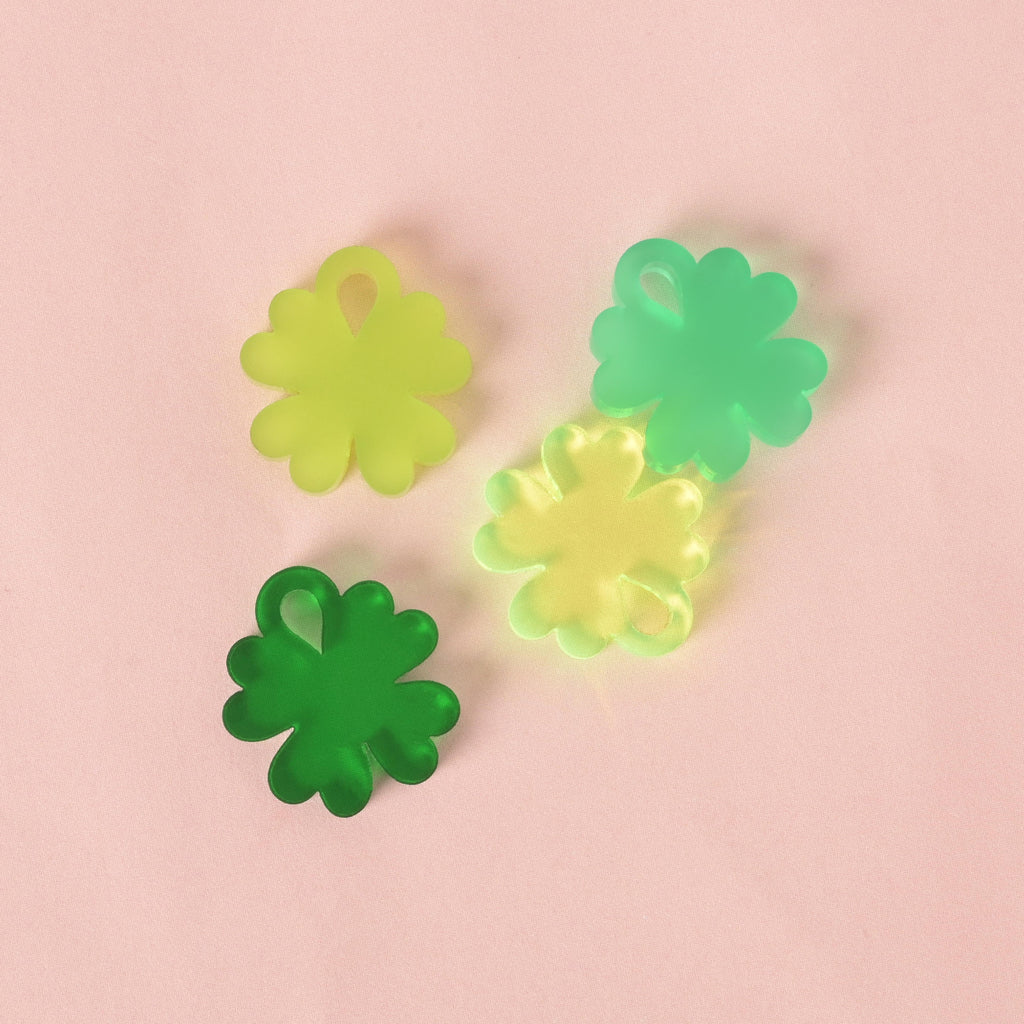 A group of four translucent, four-leaf clover-shaped charms. They are in various shades of green, including lime green, bright green, and a lighter yellow-green. The charms are arranged on a solid pink background.
