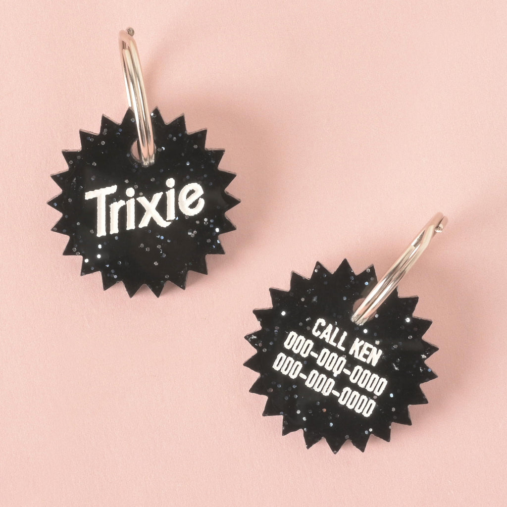 A product photo showing two sides of the same black glittery pet tag with a unique starburst shape. The tag on the left shows the front with the name "Trixie." The tag on the right shows the back, which is custom engraved with the words "CALL KEN" and two lines for phone numbers. Both tags have a silver split key ring attached.